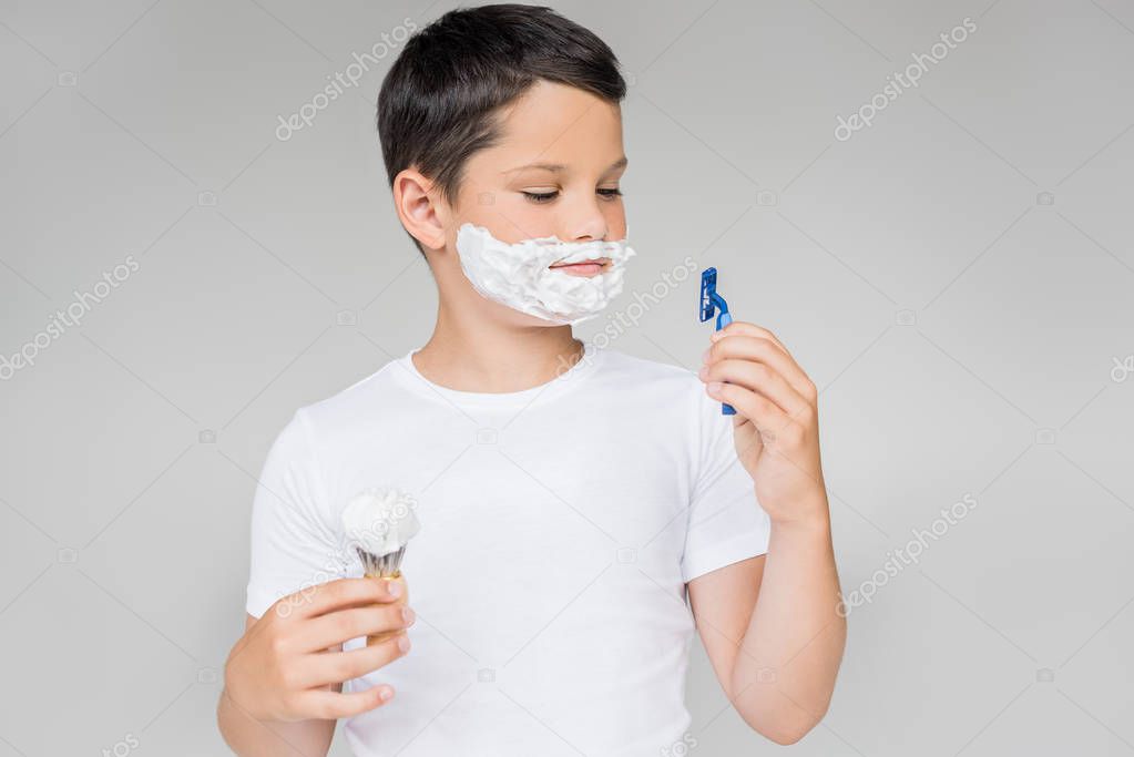 portrait of preteen boy with razor, brush shaving foam on face isolated on grey