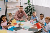 muž učitel a mnohonárodnostní předškolní děti sedí u stolu s barevné papíry v učebně