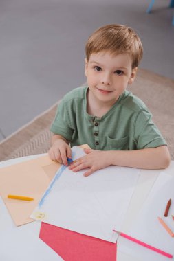 high angle view of preschooler boy sitting at table with paper and colorful pencils for drawing in classroom clipart