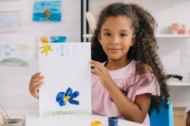 portrait of african american kid showing colorful picture in hands while sitting at table in room clipart