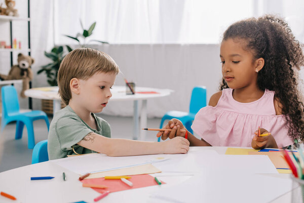 portrait of multicultural preschoolers at table with papers and pencils in classroom