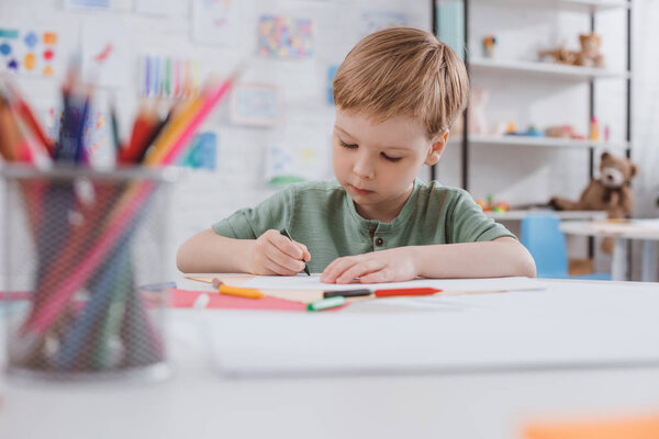 portrait of preschooler drawing picture with pencils at table in classroom