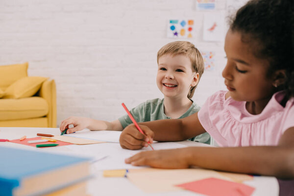 selective focus of multicultural preschoolers at table with papers and pencils in classroom