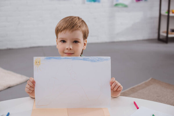 portrait of cute boy showing picture in hands at table in classroom