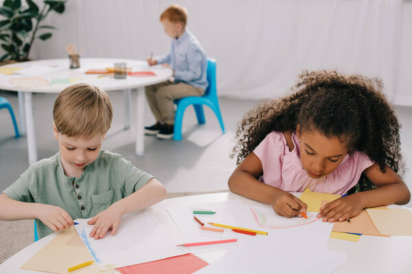 multicultural preschoolers drawing pictures with pencils in classroom
