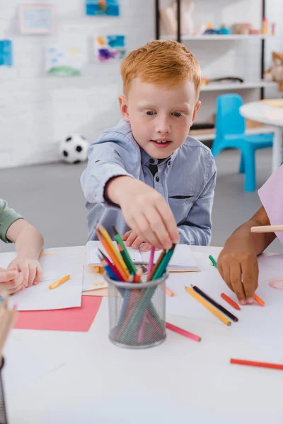 多文化共生自宅教室でクラスメイトとテーブルで鉛筆を取って赤髪少年の選択と集中  — 無料ストックフォト
