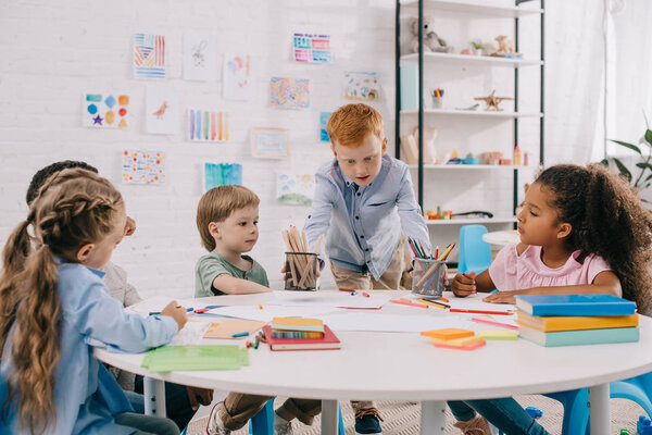 interracial kids sitting at table with papers and paints for drawing in classroom