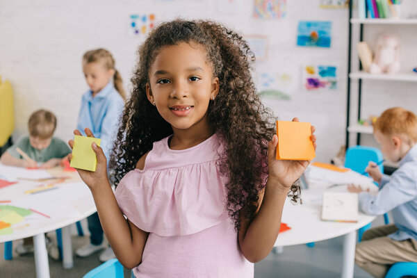 selective focus of cute african american kid with empty notes looking at camera with classmates behind in classroom