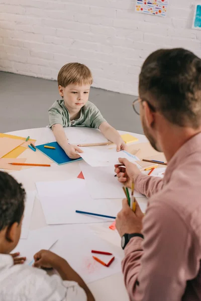 先生と教室でテーブルにカラフルな鉛筆で絵を描く多民族の少年の選択と集中  — 無料ストックフォト