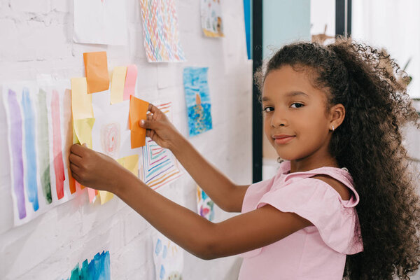side view of african american child looking at camera while hanging colorful picture on white wall in room