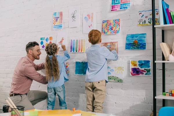 Smiling Teacher Helping Little Preschoolers Hang Colorful Pictures Wall Classroom — Stock Photo, Image