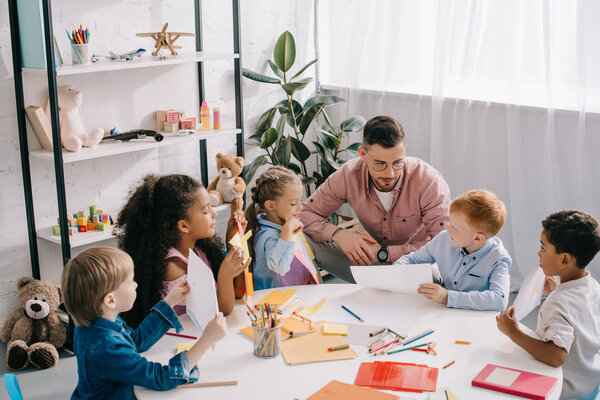 teacher and interracial preschoolers at table with paints and papers in classroom