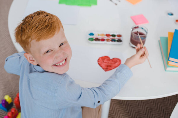 overhead view of happy red hair boy sitting at table with paints and paint brush for drawing in classroom