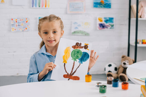 portrait of little kid showing colorful picture in hands while sitting at table in room