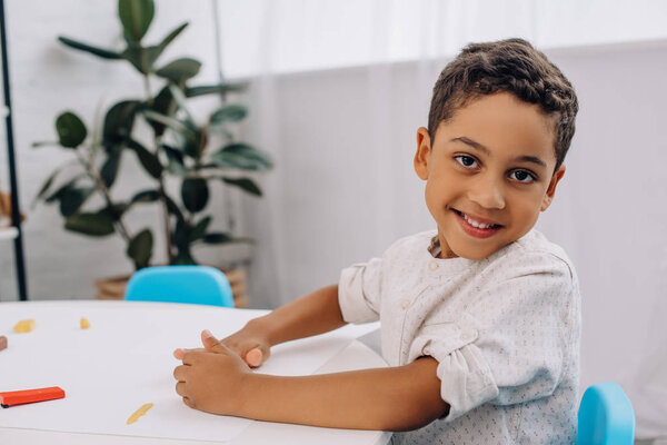 smiling african american boy with plasticine looking at camera at table in classroom