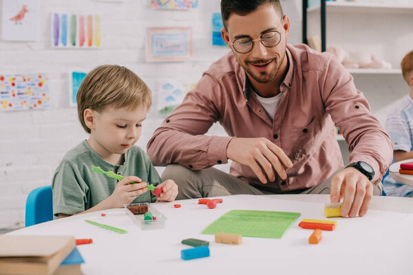 portrait of teacher and adorable preschooler with plasticine sculpturing figures at table in classroom