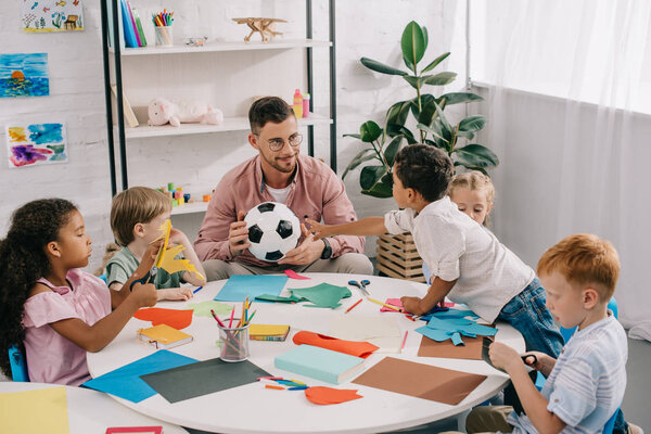 male teacher with soccer ball and multiracial preschoolers sitting at table with colorful papers in classroom