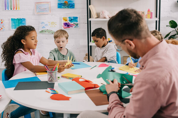teacher and multiracial preschoolers with colorful papers and scissors making paper applique in classroom 
