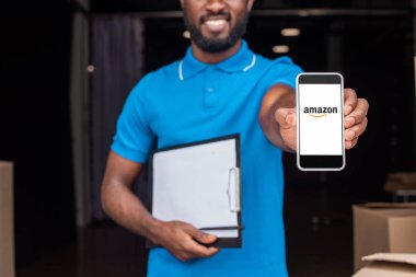 cropped image of african american delivery man showing smartphone with loaded amazon page clipart