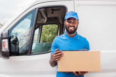 smiling african american delivery man holding cardboard box and looking at camera clipart