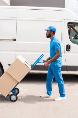 side view of african american delivery man with cart and boxes near van clipart