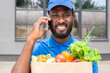 Sebzeli Kağıt torba tutarak ve smartphone ile konuşurken gülümseyen Afro-Amerikan teslimat adam