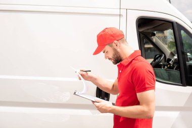 side view of caucasian delivery man looking at clipboard near van clipart