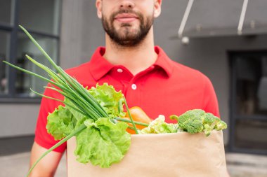partial view of delivery man in red uniform holding paper bag with fresh vegetables