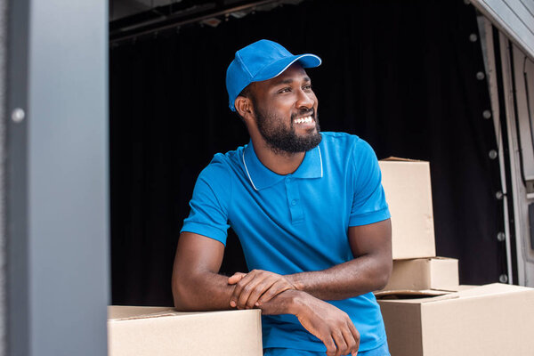 smiling african american courier leaning on boxes and looking away