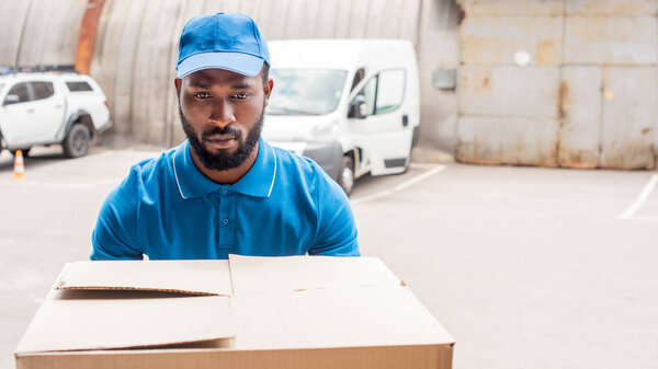 african american delivery man carrying big box with cars on background