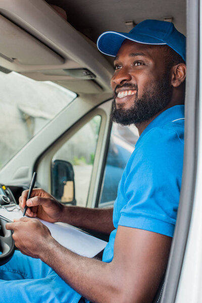smiling african american delivery man holding clipboard and pen in van 