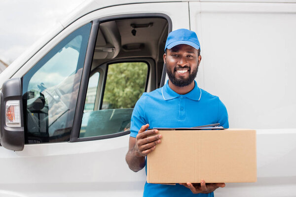smiling african american delivery man holding cardboard box and looking at camera