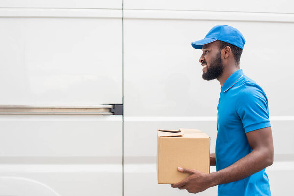 side view of smiling african american delivery man carrying cardboard box 