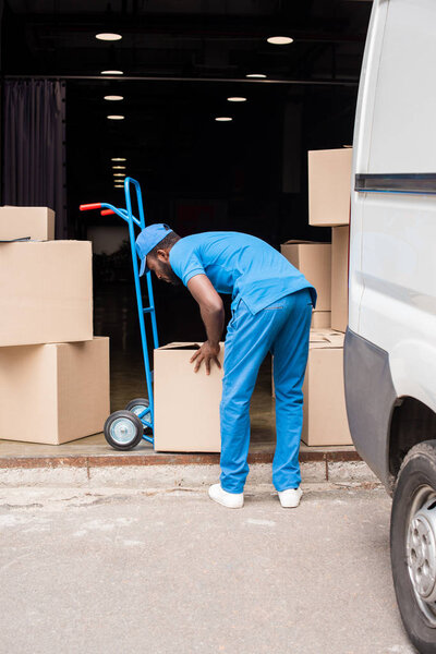 side view of african american delivery man putting boxes on cart 
