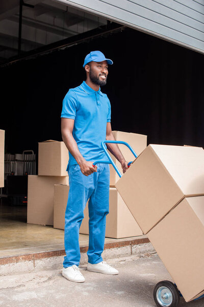 smiling african american delivery man holding cart with boxes and looking away