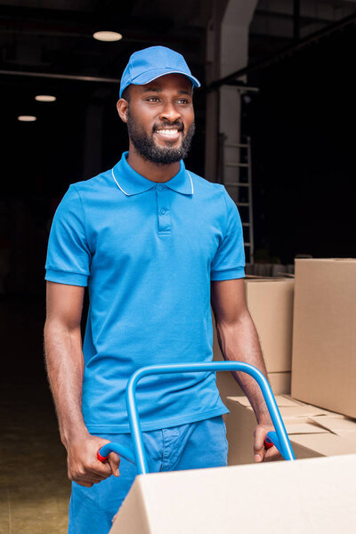 smiling african american delivery man holding cart with boxes 