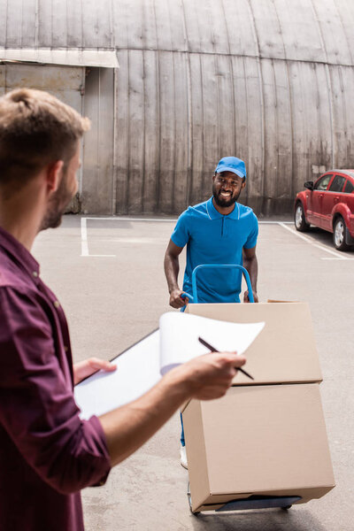 smiling african american courier with cart of boxes looking at client