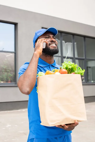 African American Delivery Man Holding Paper Bag Vegetables Talking Smartphone — Stock Photo, Image