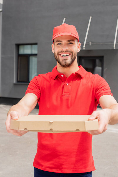 smiling caucasian delivery man holding box with pizza and looking at camera