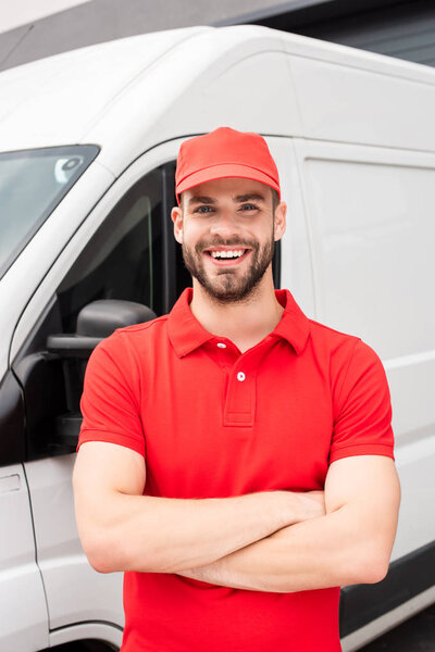 smiling caucasian delivery man standing with crossed arms and looking at camera near van