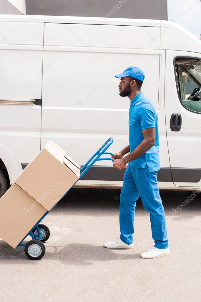 side view of african american delivery man with cart and boxes near van