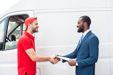 smiling delivery man and african american businessman shaking hands near van clipart