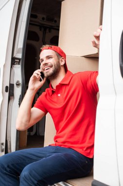smiling delivery man in red uniform talking on smartphone while resting in van clipart