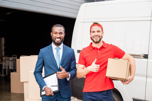 portrait of smiling multicultural client and delivery man showing thumbs up with cargo behind