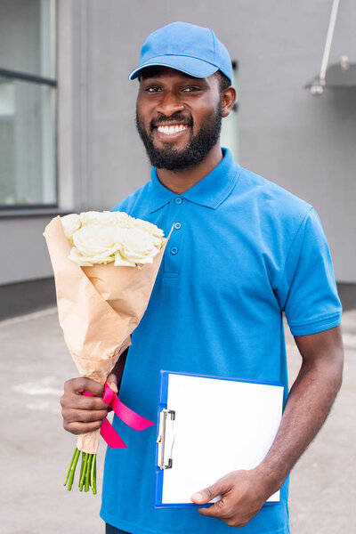 portrait of smiling african american delivery man with bouquet of flowers and cardboard in hands 