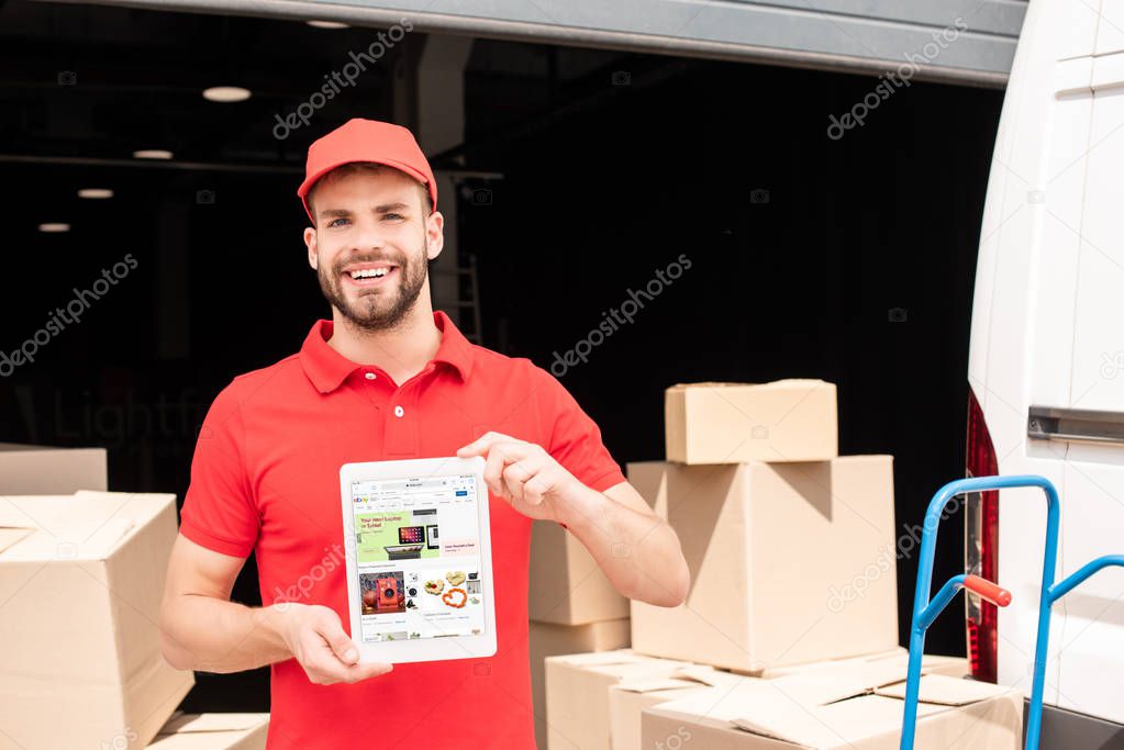 Portrait of smiling delivery man showing tablet with ebay website on screen in hands with cargo behind