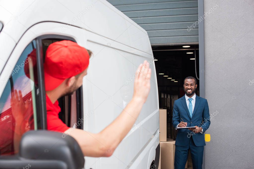 delivery man in van waving to smiling african american businessman with notepad