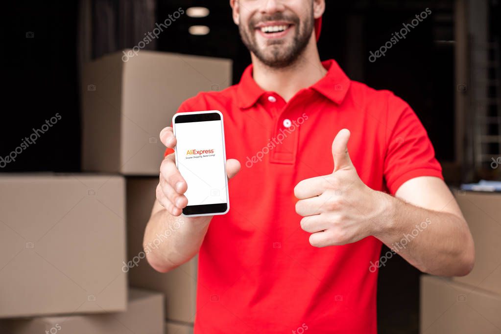 Partial view of cheerful delivery man with smartphone with aliexpress logo on screen showing thumb up