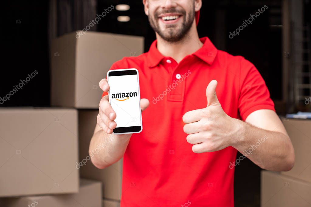 Partial view of cheerful delivery man with smartphone with amazon logo on screen showing thumb up