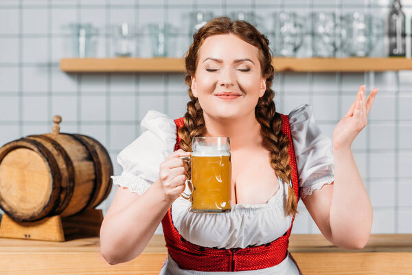 oktoberfest bartender with cloesed eyes in traditional bavarian dress enjoying smell of light beer near bar counter